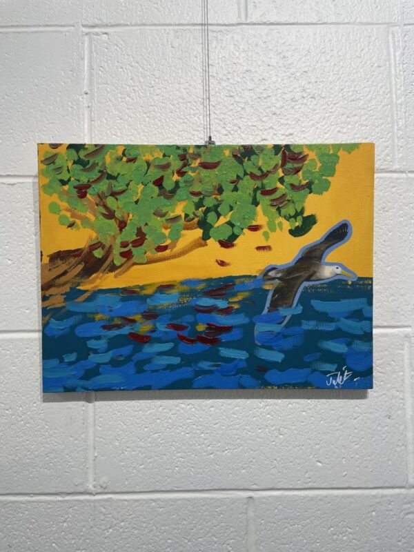 A gull flying with a blue ocean, a yellow sky and a brown and green tree.