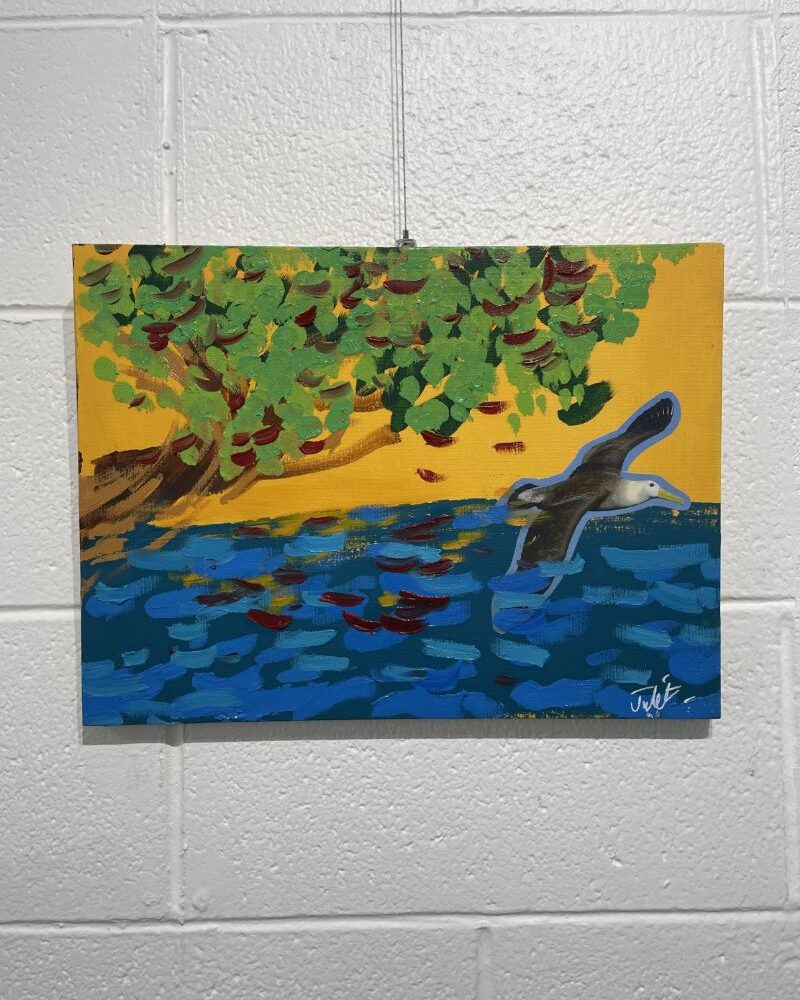 A gull flying with a blue ocean, a yellow sky and a brown and green tree.